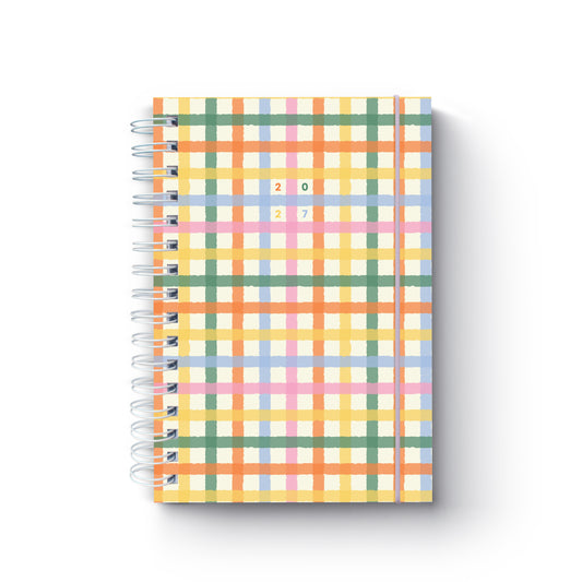 Agenda diária de 2027 Mosa com padrão axadrezado colorido em tons de amarelo, laranja, verde, azul e rosa, encadernação em espiral branca e fecho elástico lilás, sobre um fundo neutro.